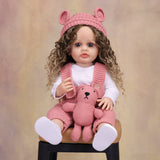 KEIUMI Sweetpea reborn toddler doll with hazel eyes and pink knitted overalls.