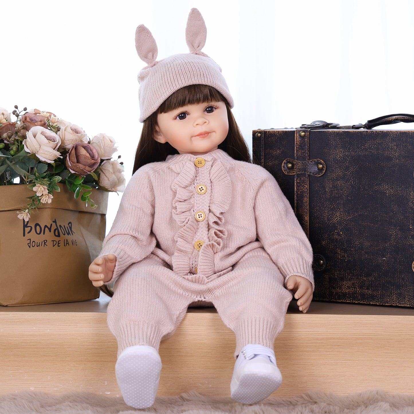 Poppy silicone reborn toddler doll in pink bunny hat outfit, sitting.